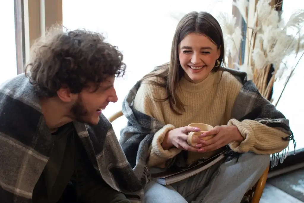 A couple smiling and talking together while wrapped in blankets, enjoying warm drinks.