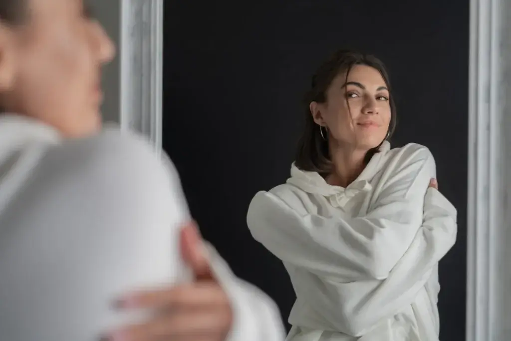 Woman smiling at her reflection while hugging herself, symbolizing self-acceptance and authenticity.